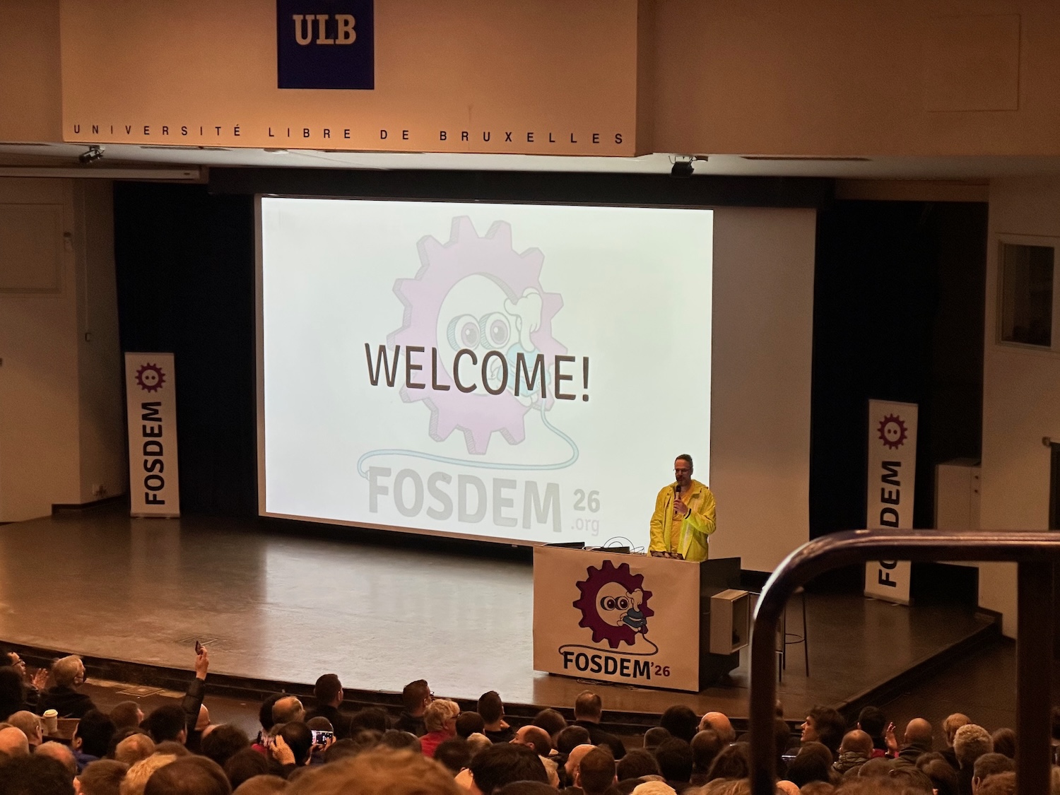 Speaker delivering the FOSDEM 2026 welcome session to a packed auditorium at ULB Brussels